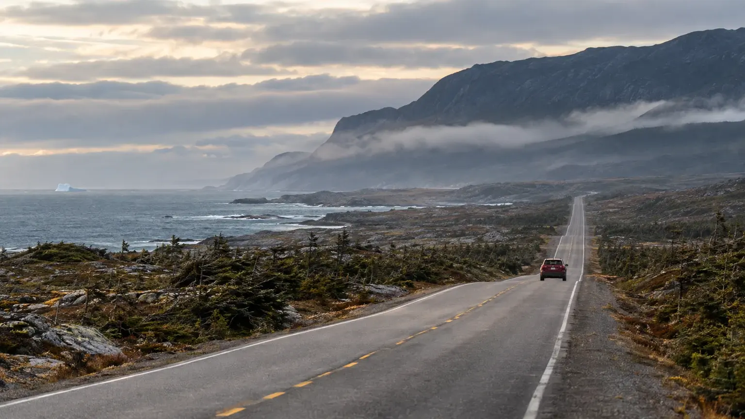 A small red car travels north on Route 430 between coastal barrens and the Gulf of St. Lawrence